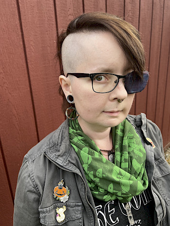 Author, a white queer individual with side-shaved auburn hair and a purple-blue shifting dupioni eyepatch, wearing a gray cotton jacket with various enamel pins and a green scarf depicting mushrooms over a black tank top which reads "The Void: No Light. No Hope. No Problem." Their jewelry includes stretched lobes with gray silicone eyelets, ash and brass coil earrings, a multi-layered septum ring, and under their scarf: leather, metal and crystal necklaces. They stand before a red barn-slatted wall. They are looking into the camera with a slight smile and lifted eyebrows.