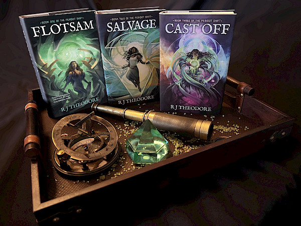 In a leather line wooden tray, various ship’s instruments sit in a scatter of peridot chips before the entries in the Peridot Shift novel trilogy: Flotsam on the far left, Salvage in the middle, and Cast Off on the right.