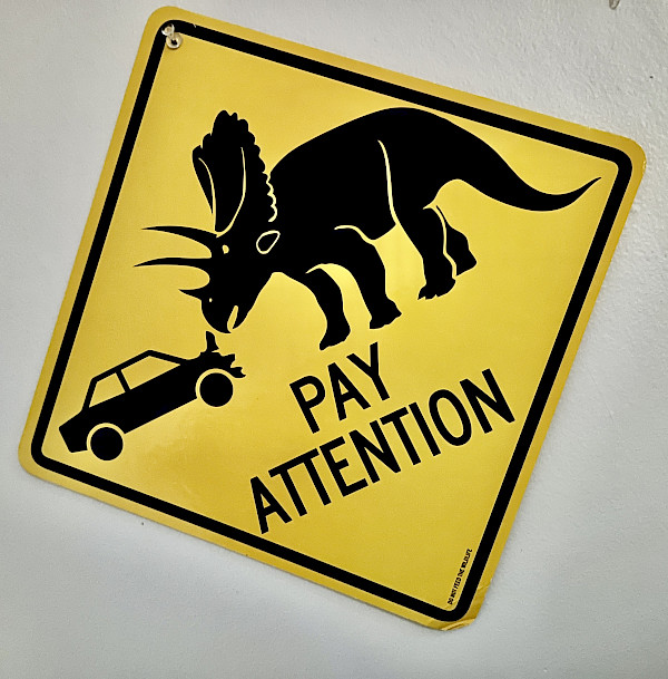 A yellow traffic warning sign that displays a car which has impacted into a triceratops with the text "PAY ATTENTION"